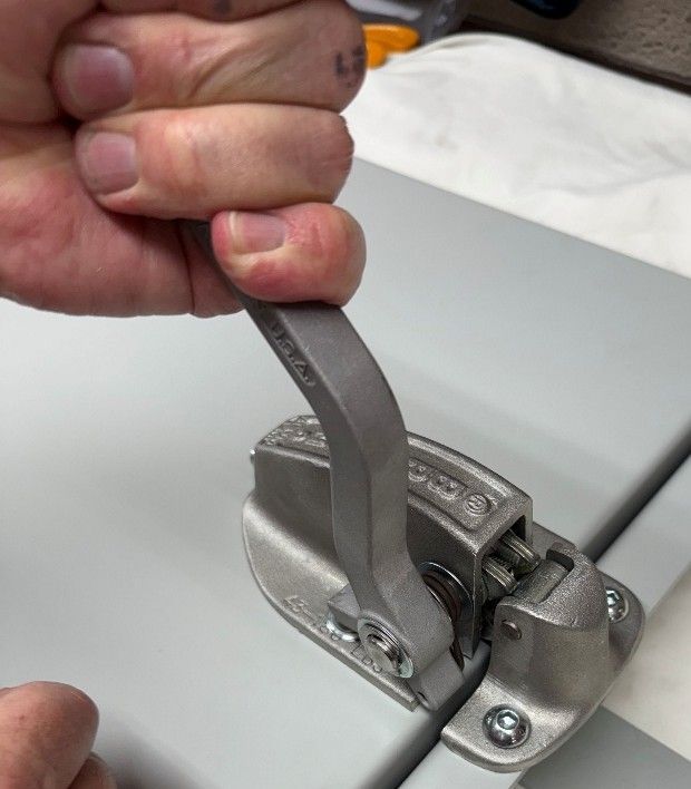 A hand operates a metal cam-style latch attached to a gray industrial panel.