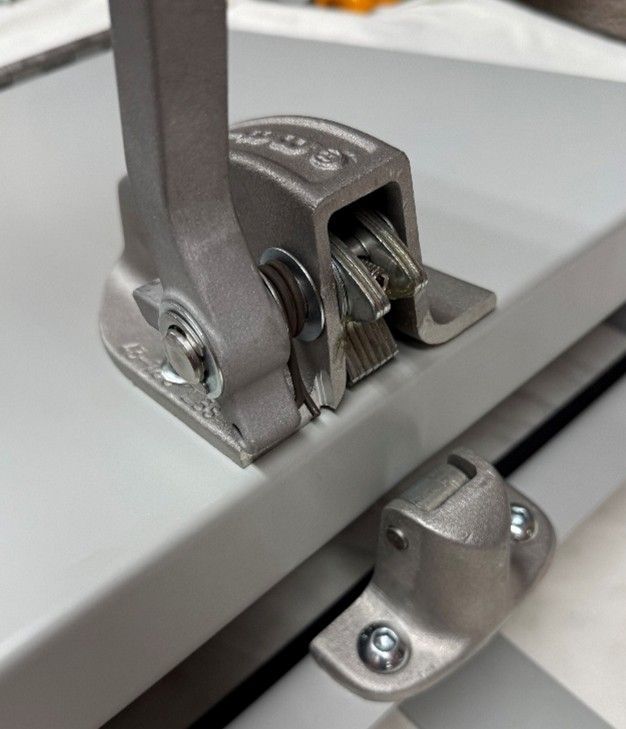A gray metal latch mechanism with a vertical handle mounted on a flat surface, with a matching catch nearby.