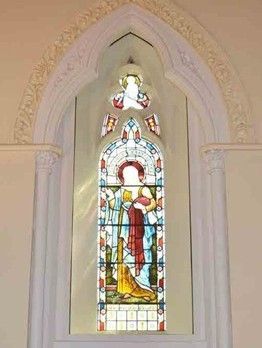 Stained glass window set in a stone arch featuring a figure draped in blue and red robes, topped with a small angel motif.