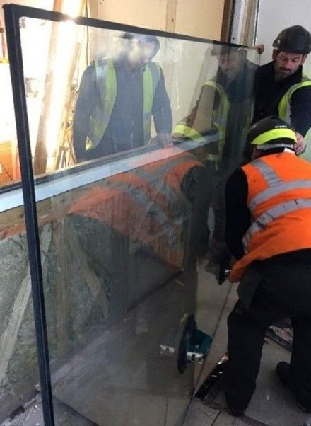 Three construction workers in high-visibility vests and helmets install a large glass pane into a wall frame.