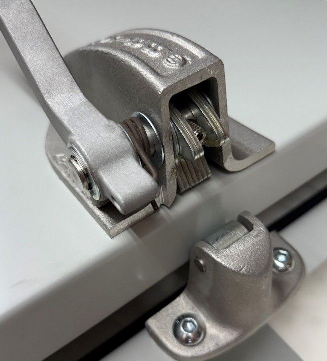 A close-up view of a metal toggle latch assembly on a light gray surface, with the locking arm partially open.
