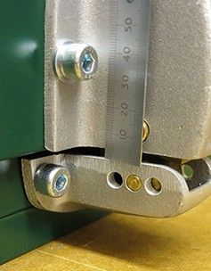 A close-up view of a metal ruler measuring the gap between a green panel and a mounted metal bracket component.