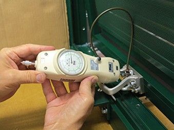 A person uses a handheld mechanical tension gauge to measure the tension on a metal cable attached to a green frame.