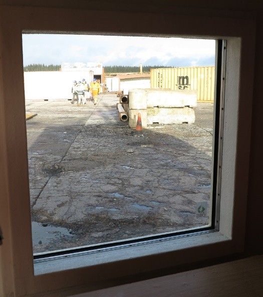A view through a window overlooking a gravel construction site with two people, shipping containers, and concrete blocks.