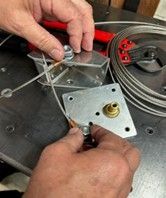 Hands assemble metal hardware and steel cable on a dark surface, with a coil of wire and red handled tools nearby.