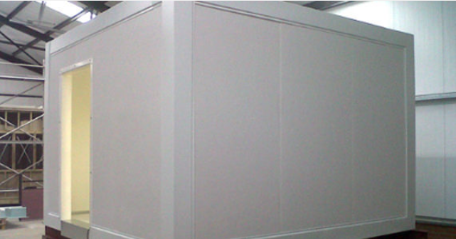 A white, modular, box-shaped structure with a door opening, located inside a workshop or warehouse setting.