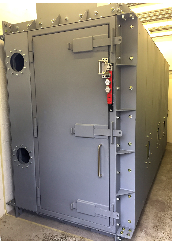 A grey, industrial walk-in enclosure with a heavy-duty hinged door, reinforced steel frame, and circular portal windows.