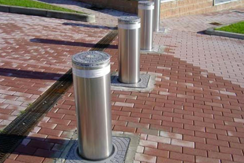 Three retractable stainless steel security bollards are standing upright on a paved, red-brick walkway.