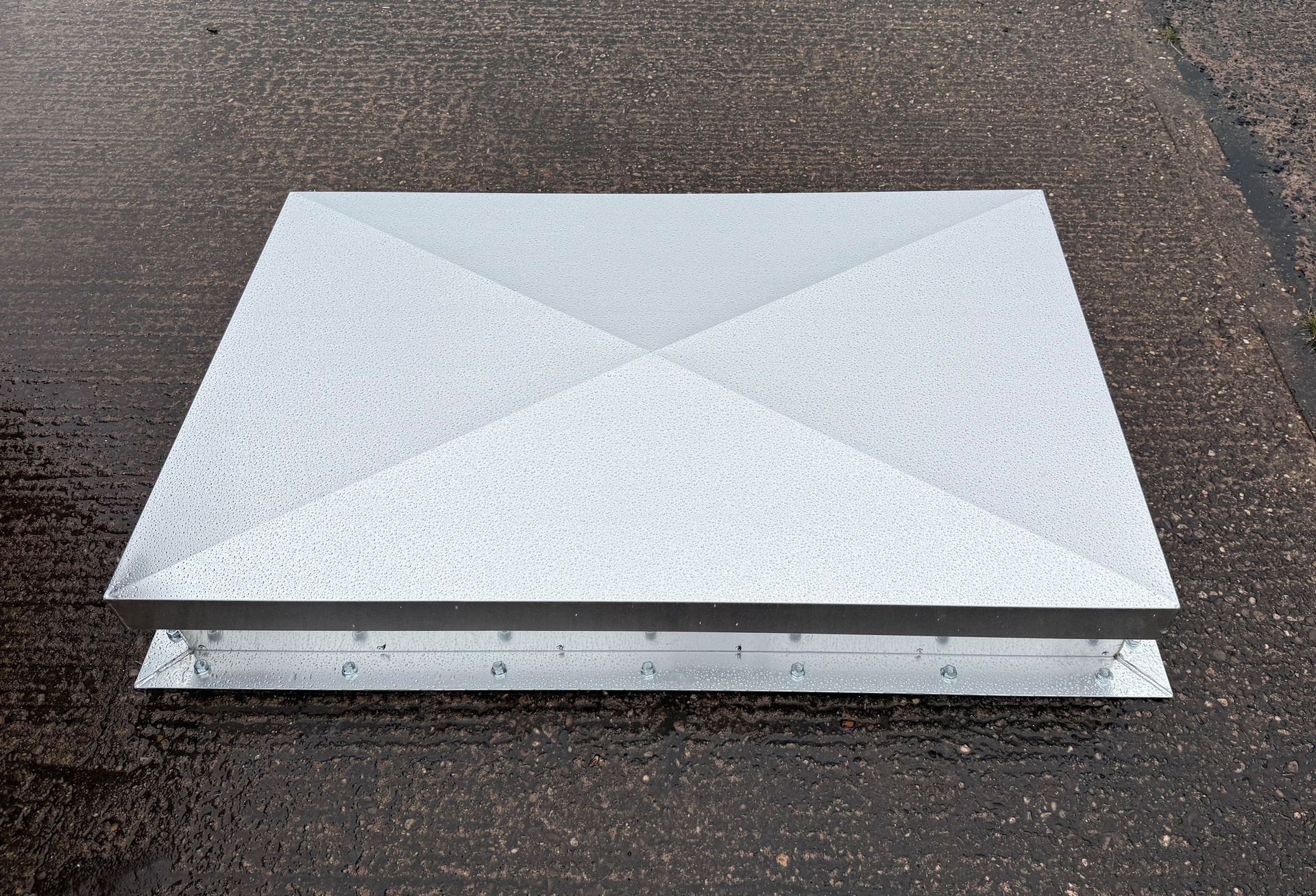 A square, silver metal roof vent cap with a four-sided sloped top, resting on a dark, wet asphalt surface.
