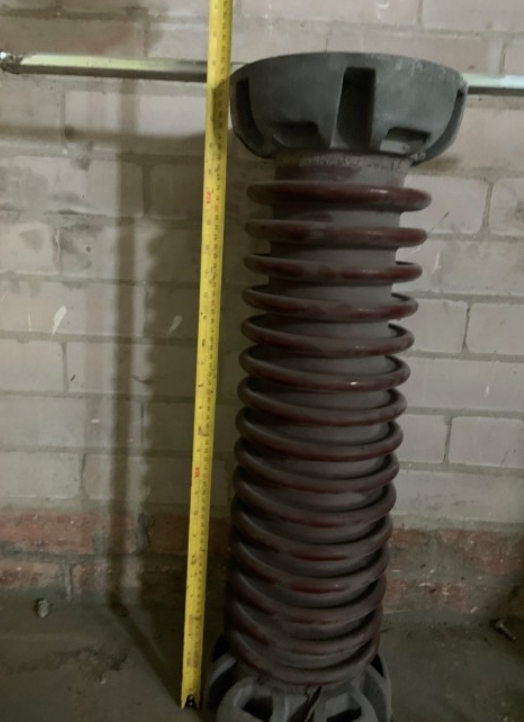 A tall, brown, ribbed high-voltage electrical insulator stands vertically next to a yellow measuring tape against a wall.