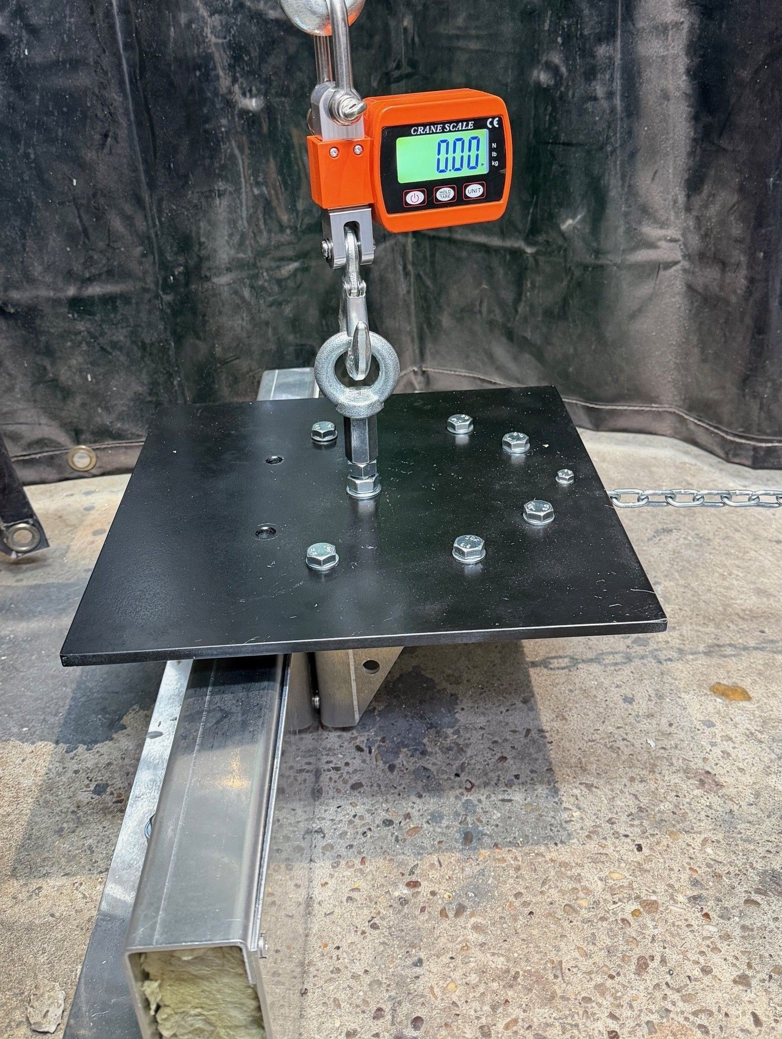 A digital crane scale hanging from a hook above a black, bolt-fastened square steel plate resting on a metal frame.