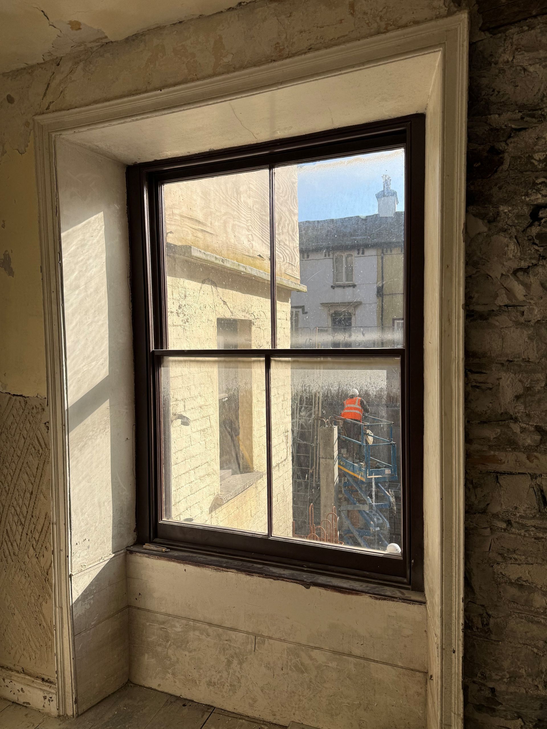 A view through a wooden-framed, four-pane window in a rustic, unfinished stone wall room looking toward another building.