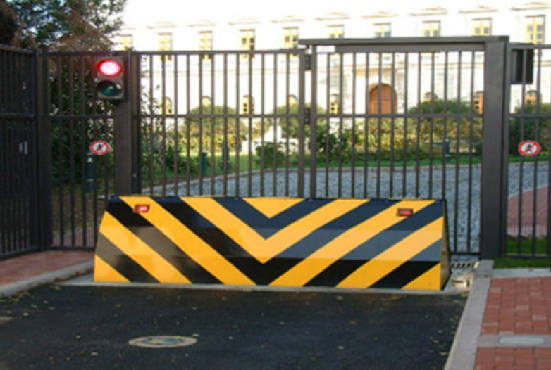 A yellow and black striped wedge-shaped rising road blocker stands deployed in front of a closed metal gate.