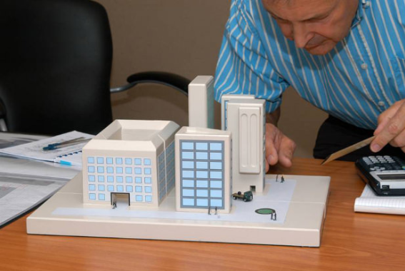 A person in a blue striped shirt leans over a table, inspecting a miniature architectural model of office buildings.