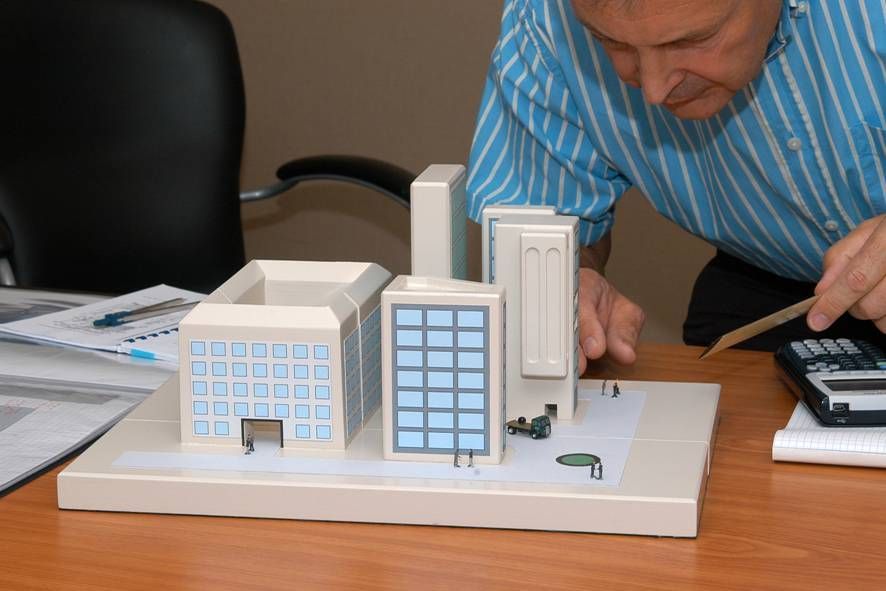 A person wearing a striped blue shirt studies an architectural model of several buildings on a wooden desk.