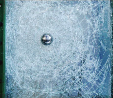 A metal ball embedded in the center of a shattered pane of glass with concentric fracture lines radiating outward.