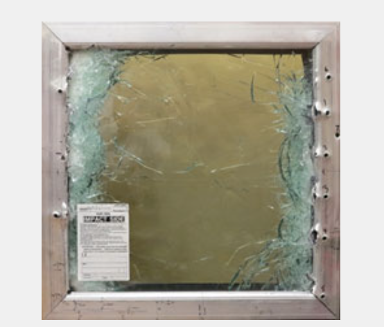 A square aluminum frame holds a pane of safety glass that is shattered around the edges, with a warning label visible.