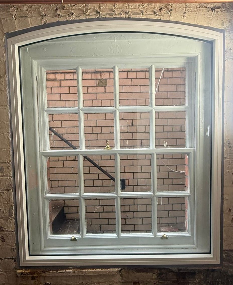 A light green, 16-pane arched window set into a rough, exposed brick wall.