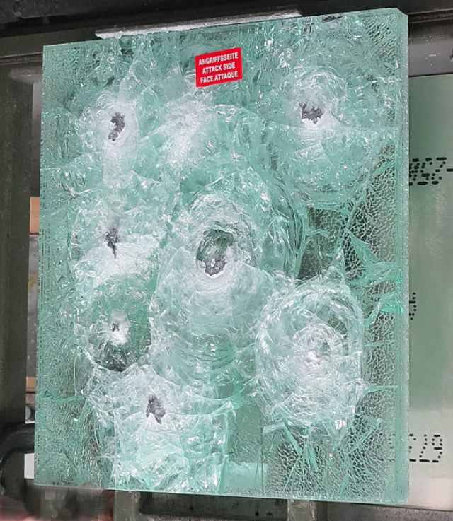 A pane of bullet-resistant glass featuring multiple impact craters and radial cracks from ballistic testing.