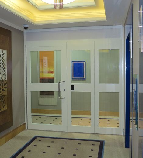 A white glass-paneled double door with a silver handle in a modern hallway with a patterned rug and recessed ceiling light.