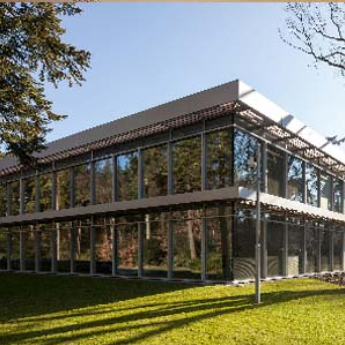 Modern two-story building with a glass facade, silver framing, and external sunshades, set in a grassy, wooded area.