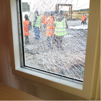 People in high-visibility safety gear stand outside a building seen through a large pane of shattered window glass.