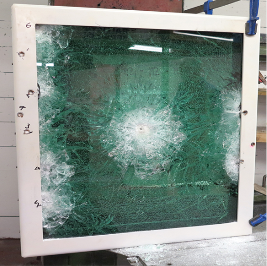 A square, white-framed panel of bullet-resistant glass featuring a prominent impact pattern and several bullet hits.