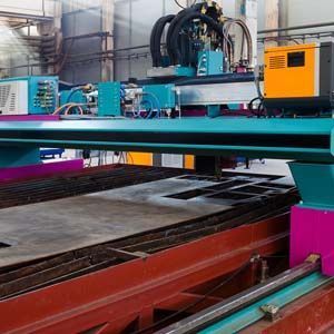 A teal and magenta industrial CNC plasma cutting machine operates on a large metal sheet in a factory setting.