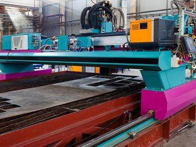 An industrial CNC plasma cutting machine operates over a large metal plate in a workshop, featuring teal, pink, and yellow.