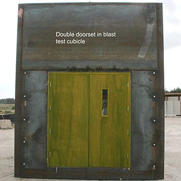 A green double-doored blast-test cubicle with a small window, set within a heavy industrial steel frame.