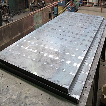 A large, thick metal plate with visible spot-welded markings sits on a workbench in a metal fabrication workshop.