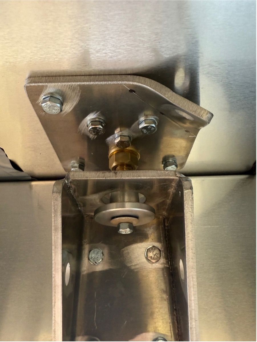 Close-up of a metal hinge assembly bolted to a brushed stainless steel surface, featuring a central brass fitting.