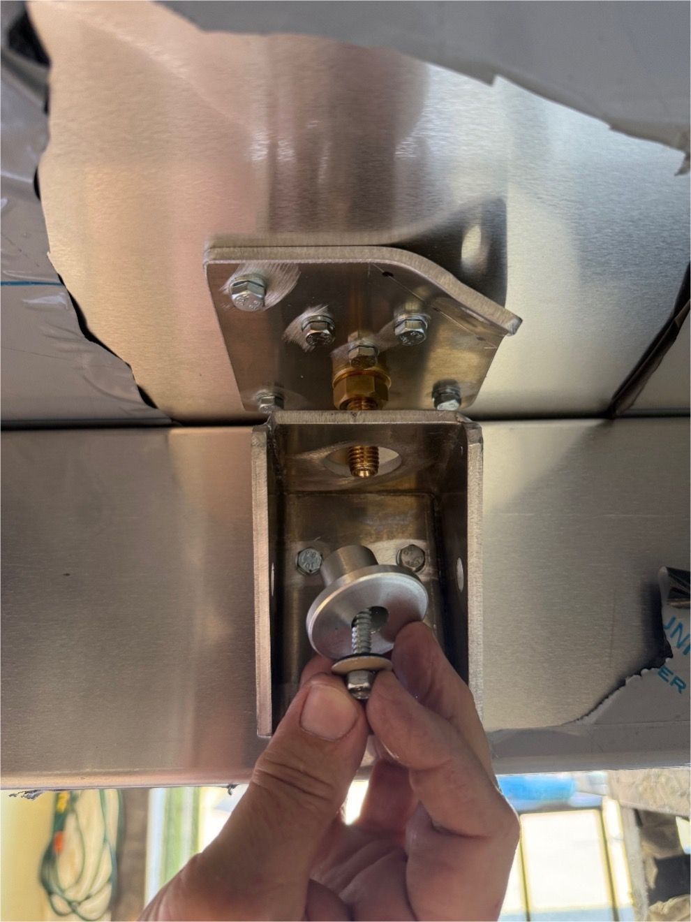 A hand aligns a metal bolt and washer through a bracket mounted on a stainless steel surface during a construction assembly.