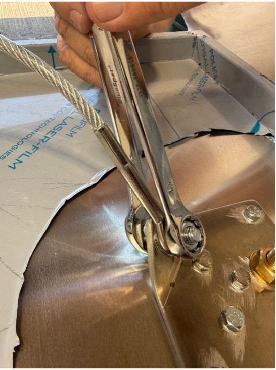 A hand uses a chrome wrench to tighten a bolt on a metal surface attached to a steel cable.