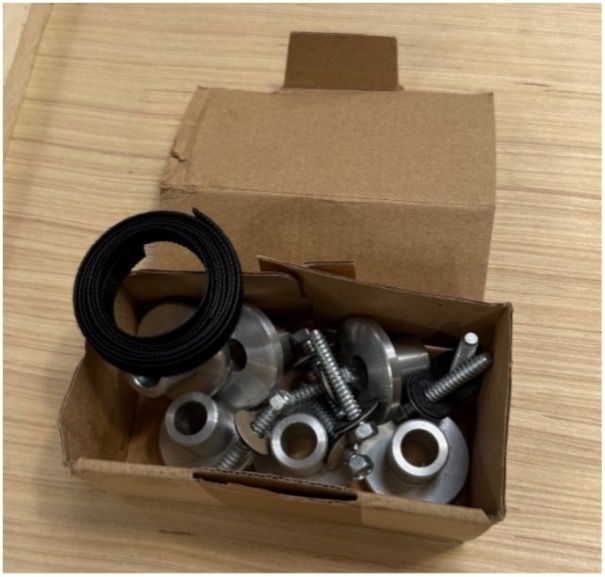 A cardboard box filled with metallic hardware fasteners, including circular spacers, screws, and a black rubber gasket.
