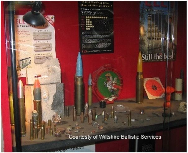 A display case containing various ammunition, bullet casings, and test targets against a red wall.