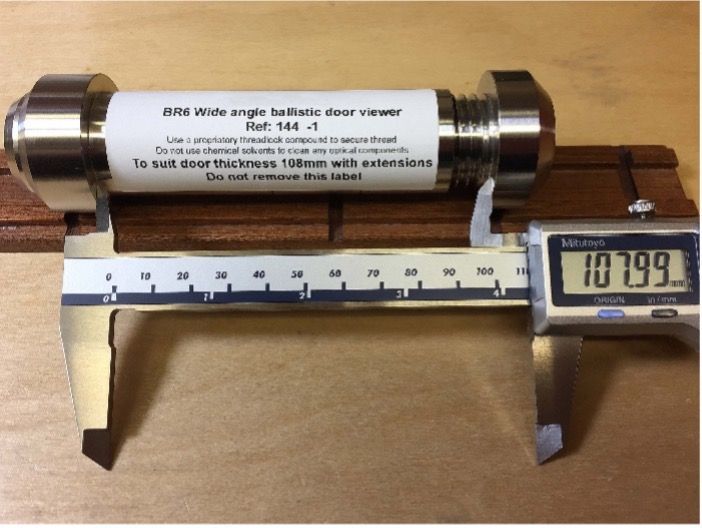A ballistic door viewer is being measured with digital calipers, which show a reading of 107.99 mm.