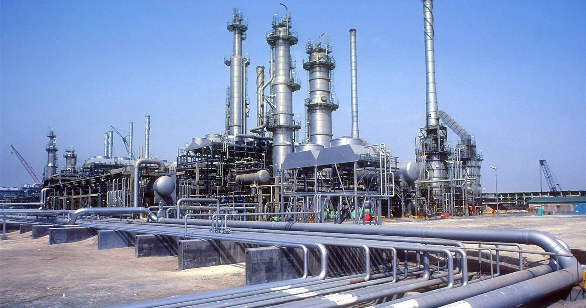 Industrial oil refinery complex with tall metallic distillation towers, extensive piping, and storage tanks under blue sky.