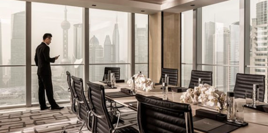 A professional in a suit stands by floor-to-ceiling windows overlooking a city skyline in a modern, furnished boardroom.