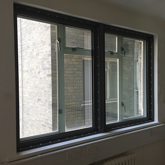 A dual-pane window with a black frame and visible fasteners installed against a white wall.