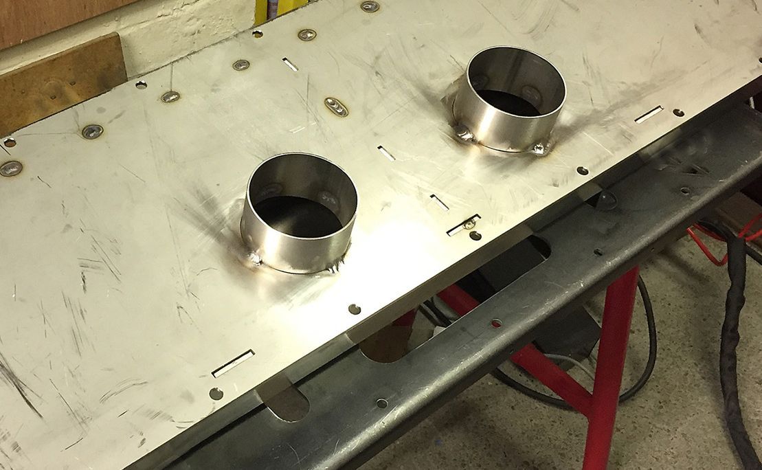 A flat metal sheet with two circular collars welded onto its surface, resting on a workshop table.