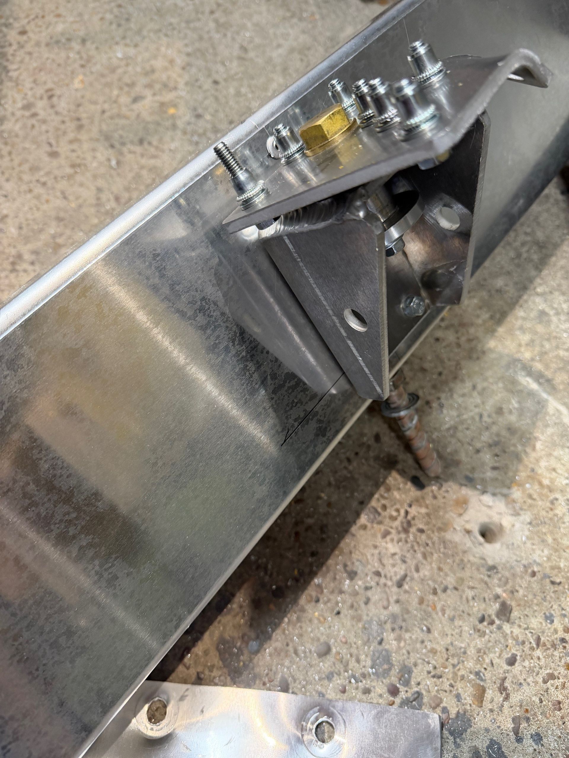 A metal mounting bracket welded to a steel beam, featuring small threaded studs and a brass-colored hex nut.