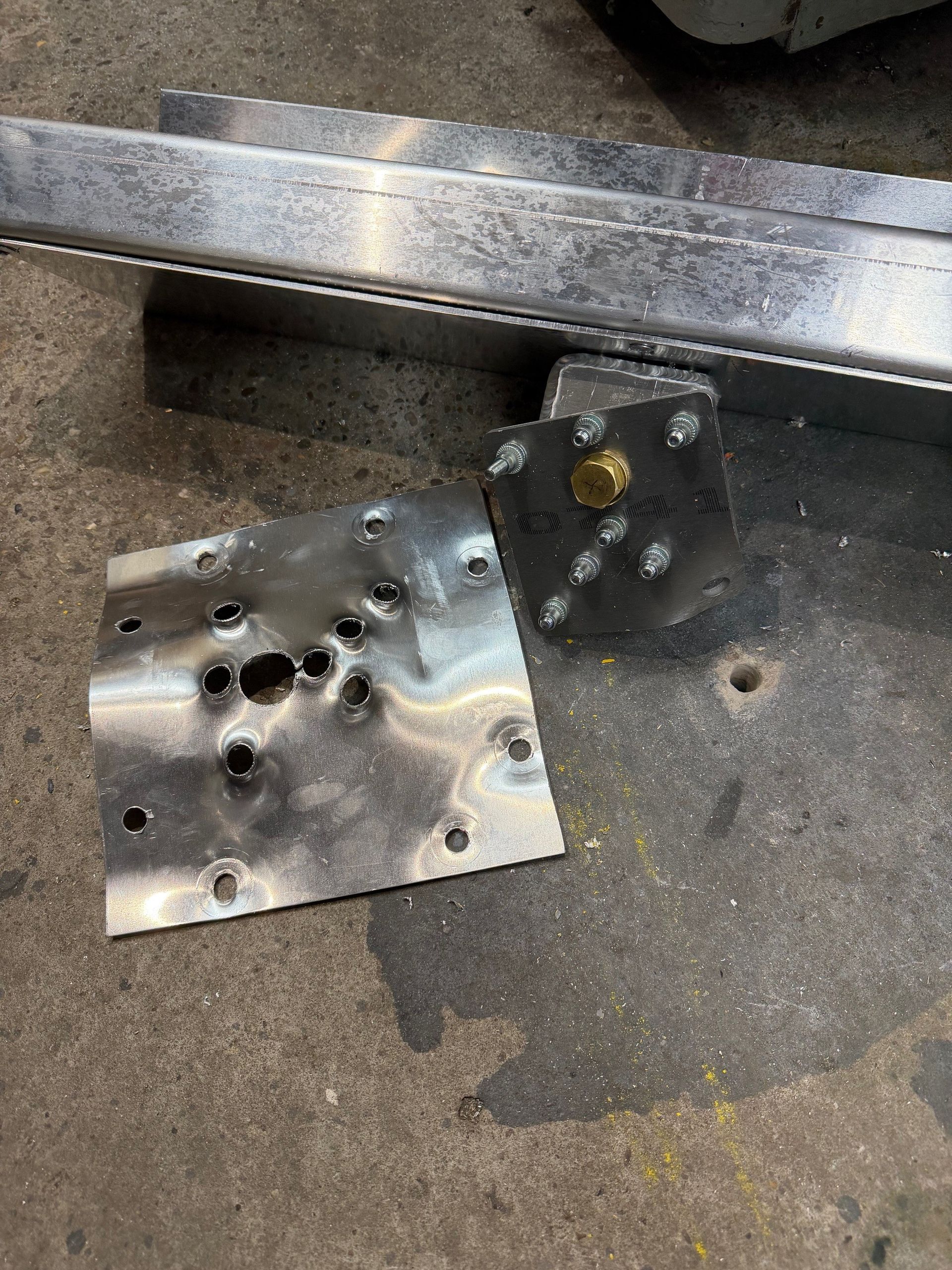 A metal bracket with multiple holes and a partially detached, bent metal plate resting on a concrete floor.