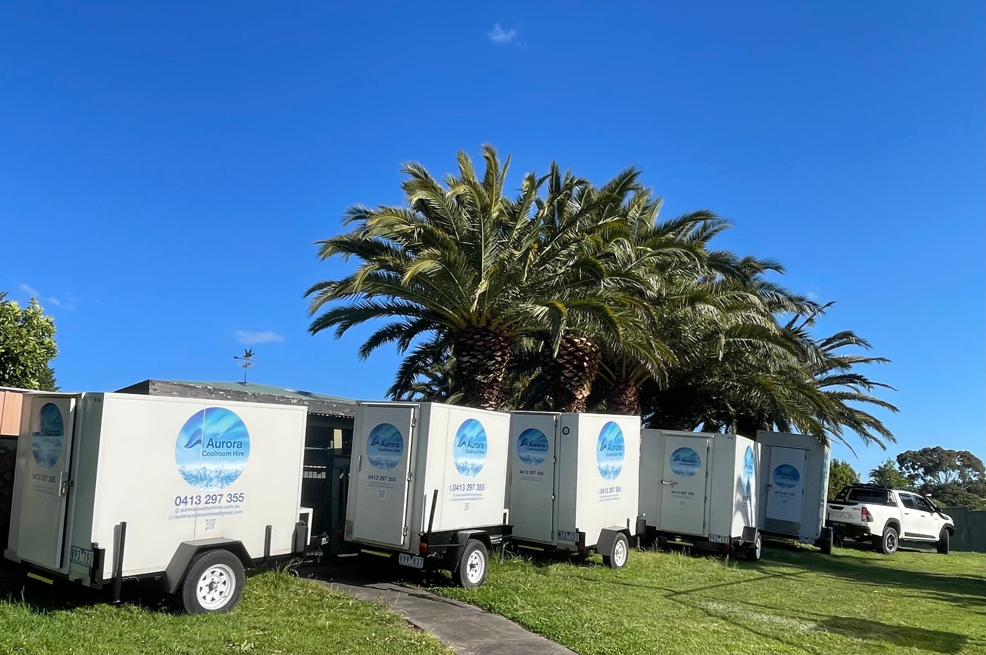 White portable toilet trailers lined up on grass, blue sky and palm trees in background.