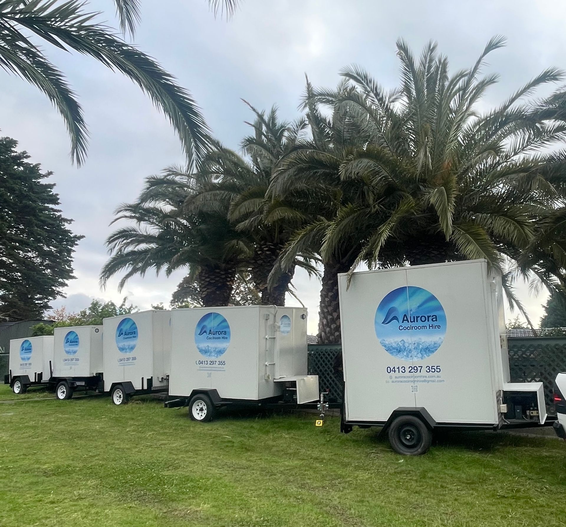 White portable restroom trailers parked on grass with palm trees in background.