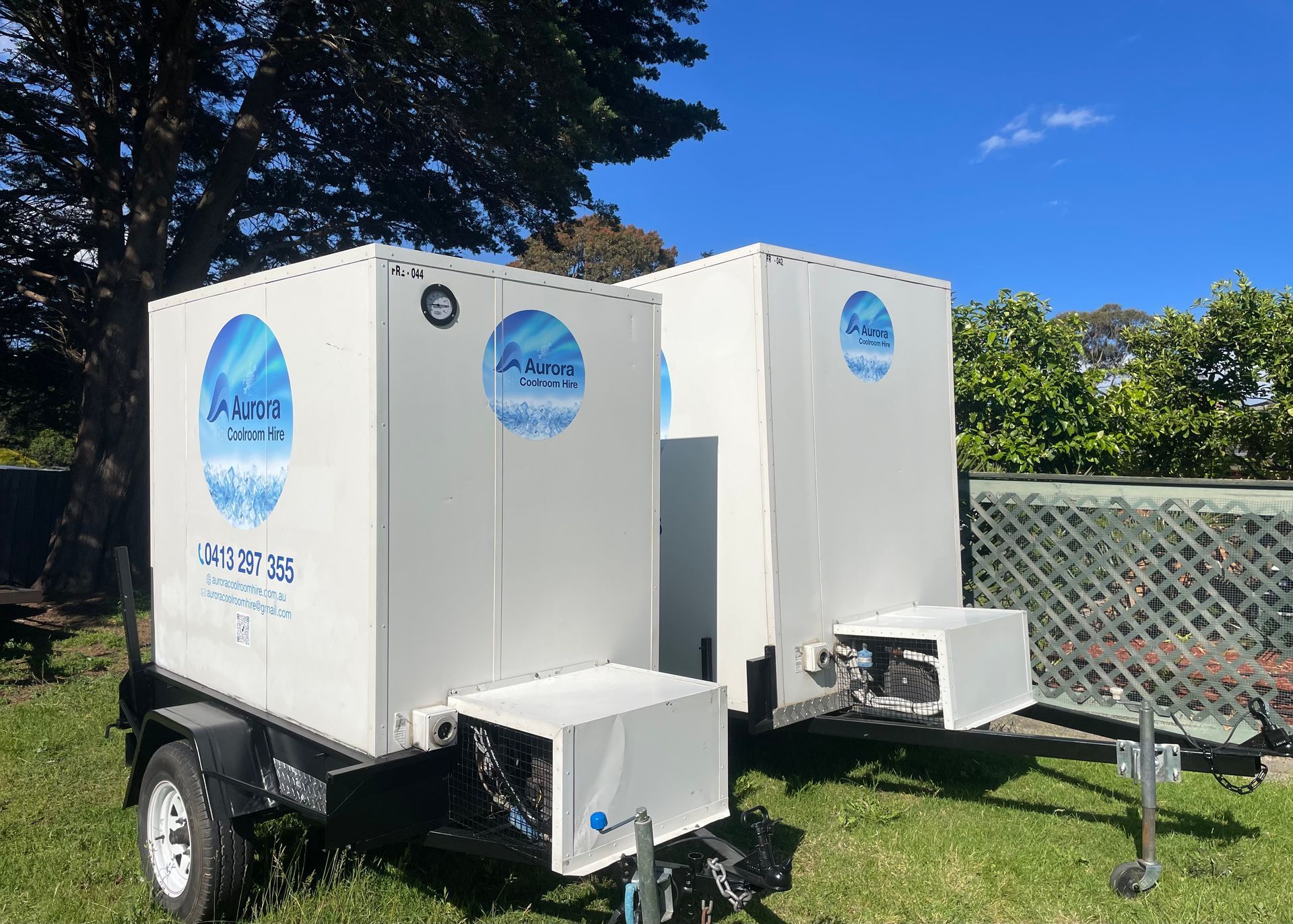 Two white mobile ice machines on trailers, with blue logos, parked on grass.