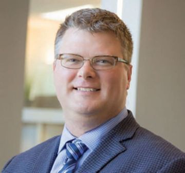 Man in glasses and blue suit smiles, office background.