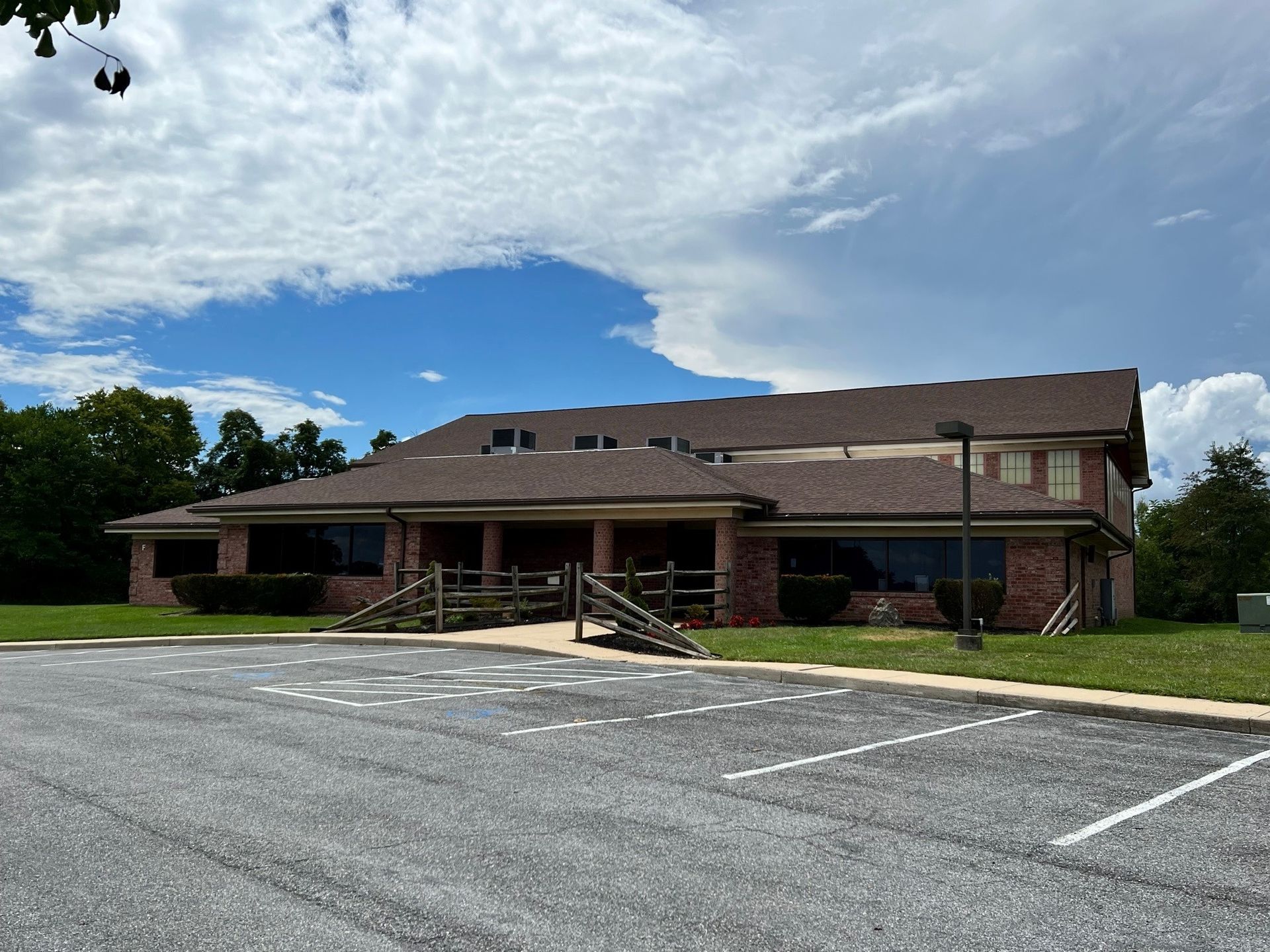 A brick building with a brown roof and large windows, in front of an empty parking lot on a sunny day.