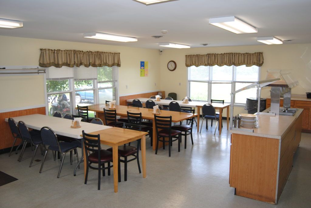 A cafeteria with tables, chairs, and a serving counter. Natural light from windows.