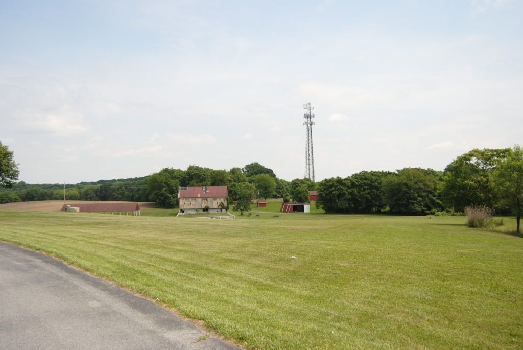Open field with buildings, trees, and a tall communication tower under a cloudy sky.